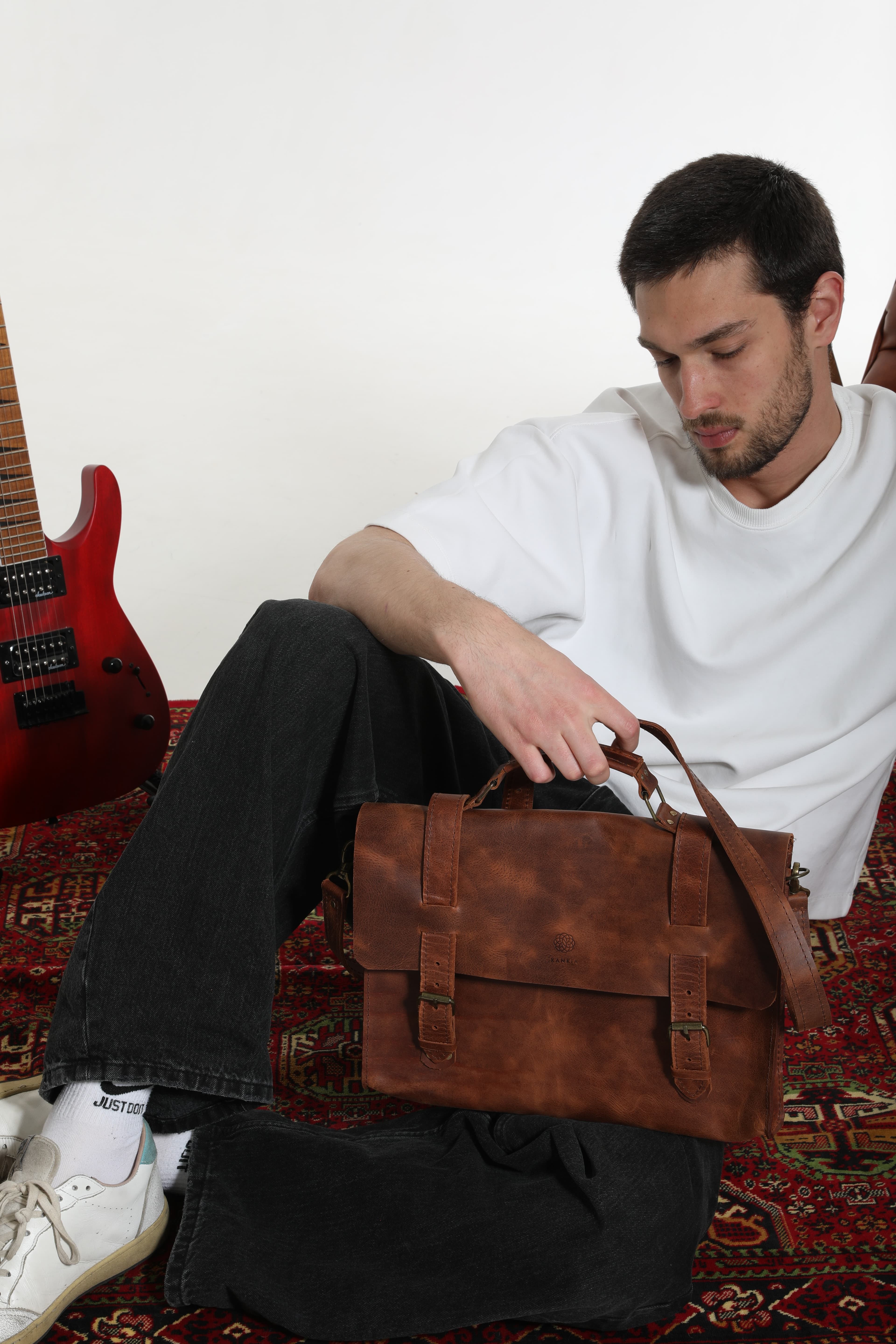 Overhead lifestyle shot — model relaxing on a colourful Georgian rug with a Kankia brown messenger bag
