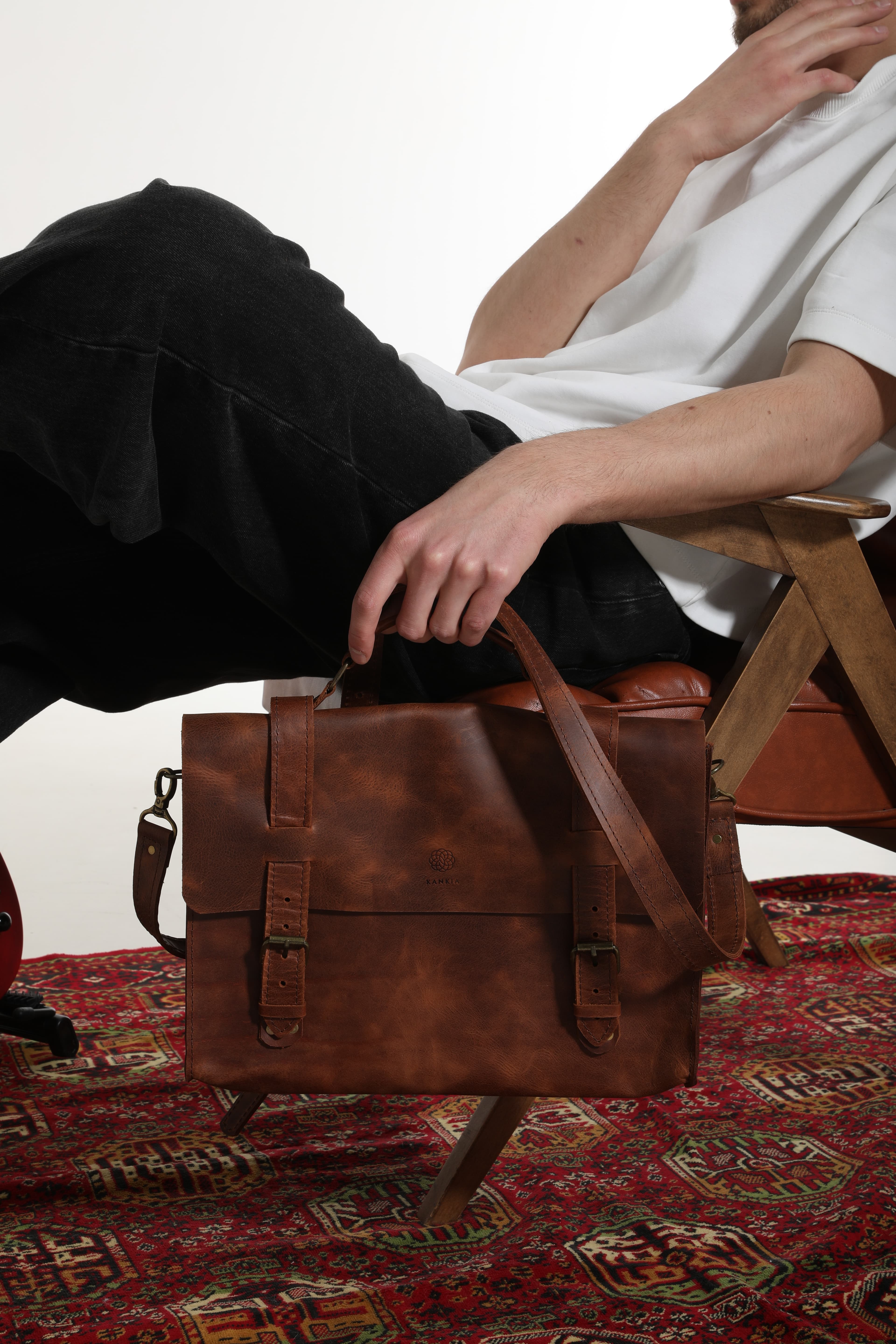 Kankia brown leather messenger bag resting on a colourful Georgian carpet, model reaching for it