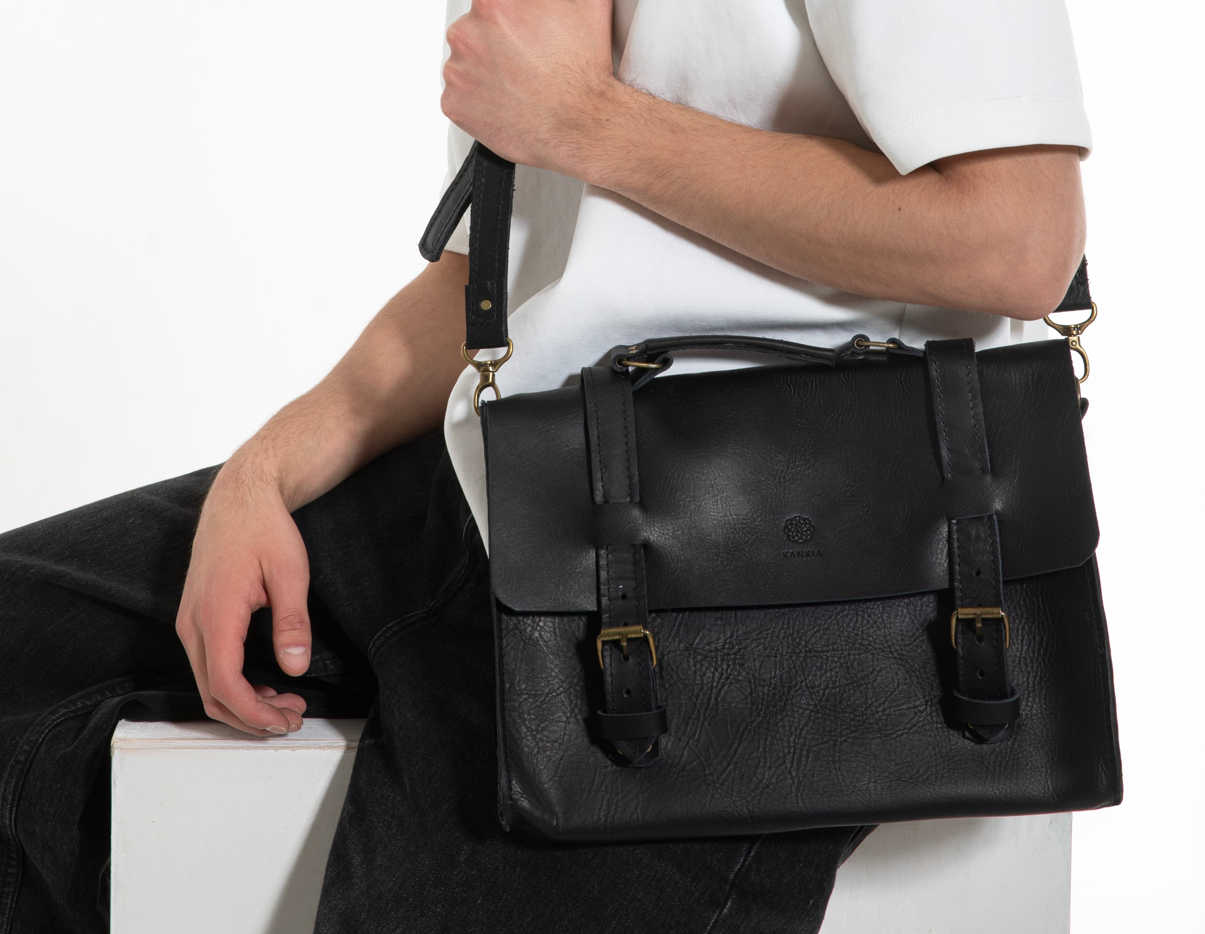 Model seated on a white block holding a Kankia black leather satchel briefcase with brass buckles